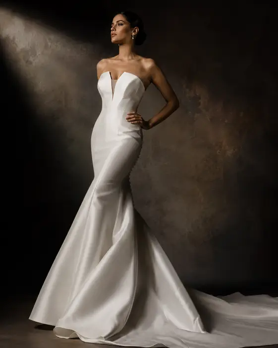 Create a dramatic editorial bridal portrait of a young woman standing tall with strong, confident posture, chin slightly raised and one hand resting on her hip. She wears a sleek modern mermaid-style wedding gown with a structured bodice and a sweeping train. Her hair is styled in a sharp, polished low bun and her makeup is bold with defined cheekbones and a soft matte lip. The camera is at a slightly low angle to emphasize elegance and height. Dramatic side lighting creates deep shadows and sharp contrast across the gown. The background is a dark, moody studio backdrop with rich gradient tones. Shot with a 50mm lens, high-contrast cinematic grading. Use aspect ratio 4:5.