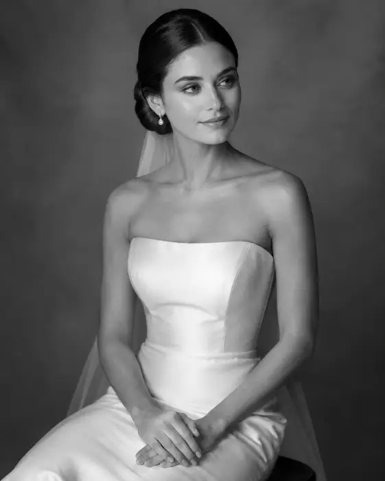 Create a timeless black and white bridal portrait of a young woman seated gracefully on a simple stool. She sits with elegant posture, hands resting softly in her lap, looking slightly off-camera with a serene, thoughtful expression. She wears a classic satin wedding gown with clean, minimal lines and a fitted bodice. Her hair is in a smooth, polished updo. The camera is at eye level, framing her from the waist up. Soft directional studio lighting creates gentle gradients of light and shadow across the gown and her face. The background is a plain, softly lit studio backdrop rendered in grayscale. Shot with an 85mm lens, rich tonal contrast. Use aspect ratio 4:5.