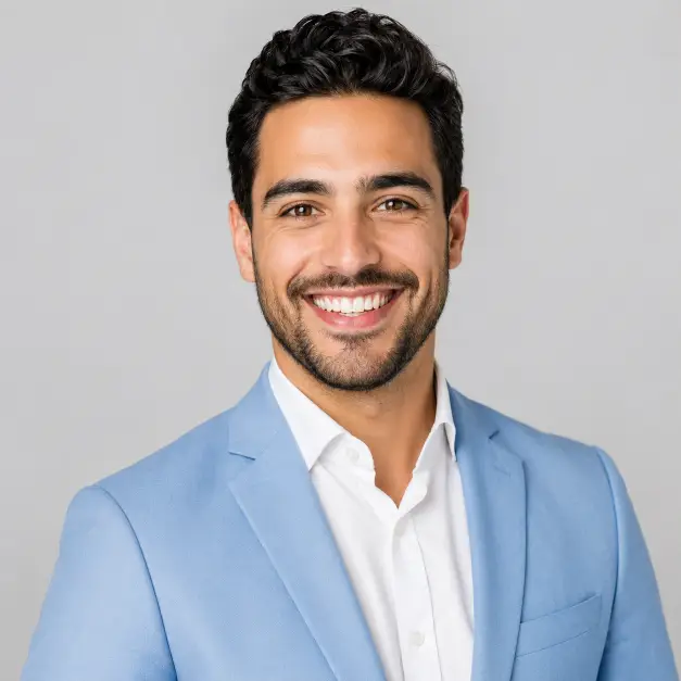 Create a bright professional headshot of a person against a clean light gray background. They are wearing a sharp light blue blazer over a white shirt, standing with confident posture and a wide, genuine smile. Their expression is energetic and warm, looking directly into the camera. The camera is at eye level with a head-and-shoulders crop. Bright, even studio lighting. The mood is upbeat, friendly, and confident. Use aspect ratio 1:1.