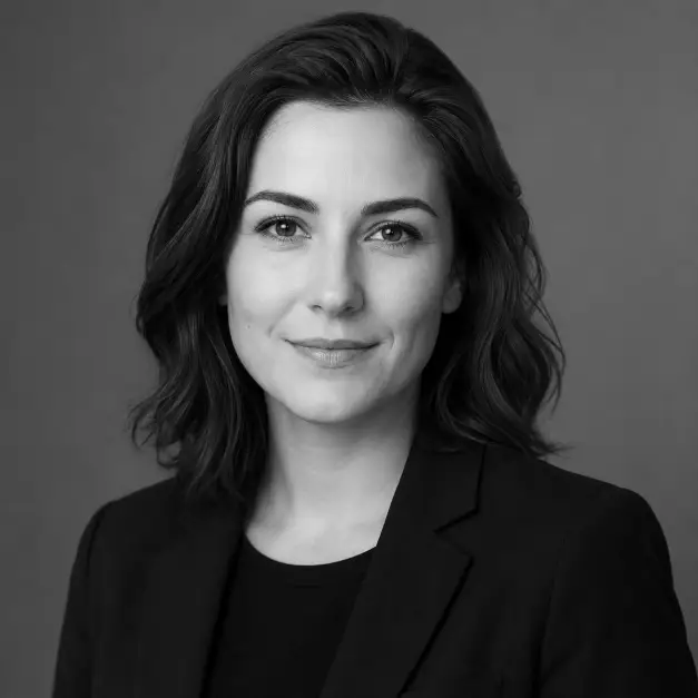 Create a timeless black-and-white professional headshot of a person against a smooth neutral background. They are wearing a simple dark blazer over a plain shirt, standing with upright posture and a calm, confident expression. Their gaze is steady and directed into the camera with a slight smile. The camera is at eye level with a head-and-shoulders crop. Soft studio lighting with gentle contrast and clean tones. The mood is classic, elegant, and professional. Use aspect ratio 1:1.