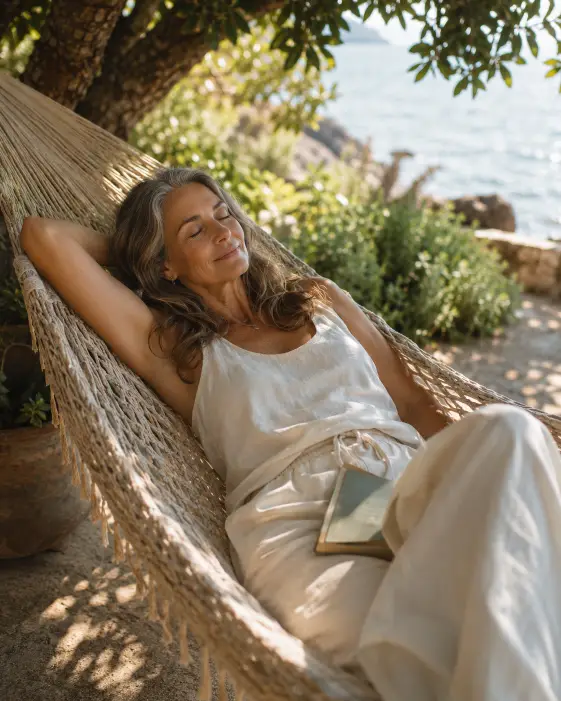 Create a photo of a woman in her early 60s lying back in a woven hammock under the shade of a tree, one arm behind her head and a book resting on her lap. She is wearing a soft cream linen set of a loose tank top and wide drawstring trousers. Her hair is down in gentle waves. Her eyes are softly closed with a peaceful expression. The background is a coastal garden with dappled light coming through the leaves and a soft blurred sea view. The camera is at a slightly high angle, looking gently down at her. Warm afternoon light with soft golden and green tones. Shot with a 35mm lens, relaxed lifestyle style. Use aspect ratio 4:5.