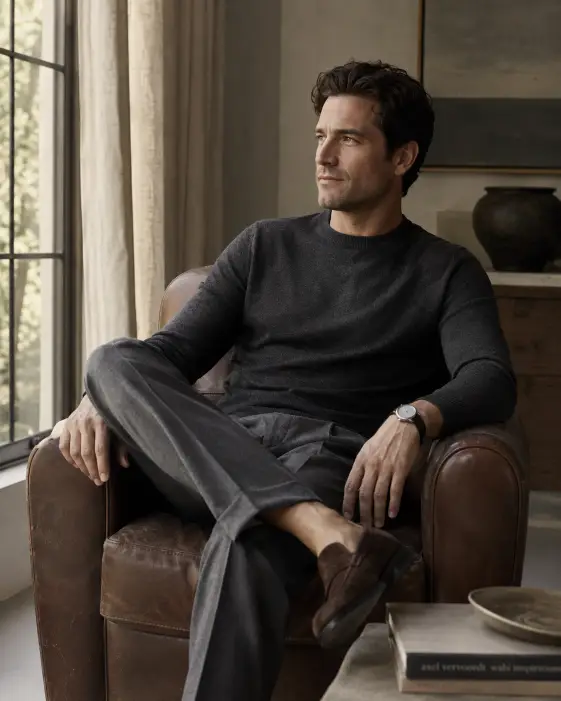 Create a photo of a man in his mid 30s sitting in a leather armchair near a tall window, leaning back with one ankle resting on his knee and a relaxed expression. He is wearing a fine-gauge charcoal merino knit, pleated grey trousers, and brown suede loafers, with a plain steel watch on his wrist. His hair is neatly styled and his expression is calm. Shoot at a slight three-quarter angle from chest height. The background is a softly blurred living room with linen curtains and warm natural light pouring in. Muted grey and beige color palette. The mood is refined and quiet. Use aspect ratio 4:5.