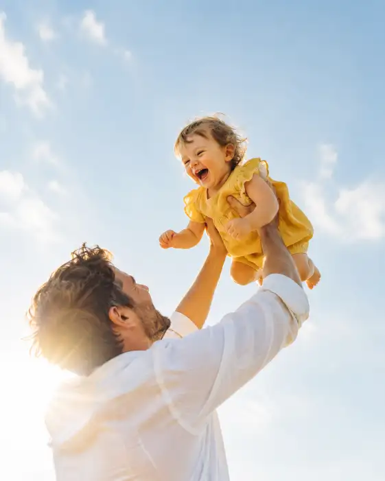 Create a photo of a father lifting his laughing toddler high into the air against a soft blue sky, both full of joy. He is wearing a relaxed white shirt and the toddler is in a yellow outfit. Shoot from a low angle looking up with bright, airy backlight and gentle sun flare. The background is a clean blurred sky with plenty of open space at the top for adding card text. The mood is uplifting, bright, and celebratory. Use aspect ratio 4:5.