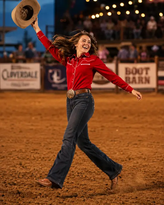 Create a dynamic photo of a young woman mid-action at a rodeo arena, one hand raised holding her hat in the air and her body twisting with energy. She is wearing a fitted red western shirt with white piping, dark bootcut jeans, a large silver belt buckle, and brown leather boots. Her hair is flying with the movement and she has a wide, joyful expression. The camera is at a slightly low angle, capturing her full body with a hint of motion blur. The background is a dusty rodeo arena with wooden fencing and blurred crowd lights. Warm, punchy tones with vivid saturation. Shot with a 35mm lens, fast shutter. Use aspect ratio 4:5.