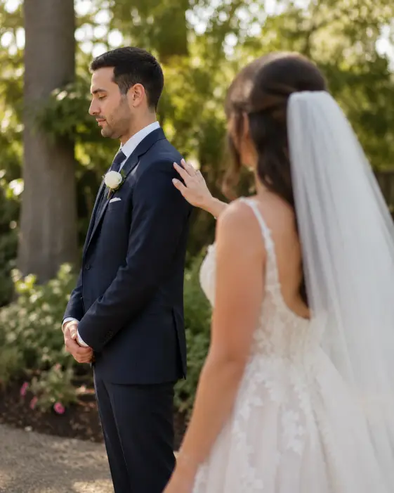 Create a candid wedding photo of a groom standing with his back to his bride as she approaches him from behind for their first look. He is wearing a fitted navy three-piece suit with a white boutonniere, his hands clasped nervously in front of him. She is in a flowing ivory A-line gown with a long veil, one hand reaching toward his shoulder. The camera is at eye level, capturing both of them in profile with the bride slightly out of focus in the foreground. Soft, warm afternoon light filters through tall trees in a garden setting with blurred greenery behind them. The mood is tender and emotional, shot with a 50mm lens and shallow depth of field. Use aspect ratio 4:5.