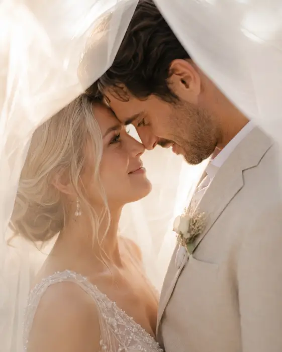 Create an intimate wedding portrait of a couple standing close together beneath the bride's sheer veil, which drapes over both of their heads like a soft frame. The bride is wearing a delicate beaded gown and is looking up at the groom, who is in a light beige linen suit with an open collar. Their foreheads are nearly touching with soft, loving expressions. The veil creates a dreamy, hazy texture around them. The camera is close at eye level with a tight composition from the shoulders up. Soft, glowing backlight comes through the veil creating an ethereal effect. Pale, romantic tones. Shot with a 50mm lens, wide aperture. Use aspect ratio 4:5.