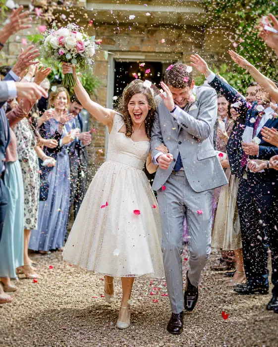 Create a joyful wedding photo of a couple walking through a tunnel of cheering guests throwing confetti and rose petals into the air. The bride is laughing with her bouquet raised, in a tea-length vintage gown, and the groom is shielding them playfully in a light gray suit. The guests on both sides are blurred with motion, arms raised. Confetti fills the air and catches the light. The camera is at eye level capturing the couple mid-stride from the front. Bright, lively daylight with vivid celebratory tones. Shot with a 28mm lens, fast shutter to freeze the confetti. Use aspect ratio 4:5.