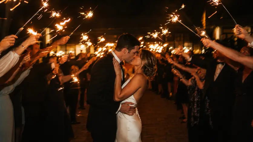 Create a magical nighttime wedding photo of a couple kissing under a shower of sparklers held by guests on both sides forming a glowing tunnel. The bride is in a satin gown and the groom in a dark suit, both lit by the warm sparks around them. The background is dark with the soft glow of the venue lights behind the guests. The sparklers create bright streaks of light and a warm glow on the couple's faces. The camera is at eye level, capturing the couple in the center of the tunnel. Dark moody background with warm glowing highlights and a cinematic feel. Shot with a 35mm lens, slightly slower shutter for light trails. Use aspect ratio 16:9.