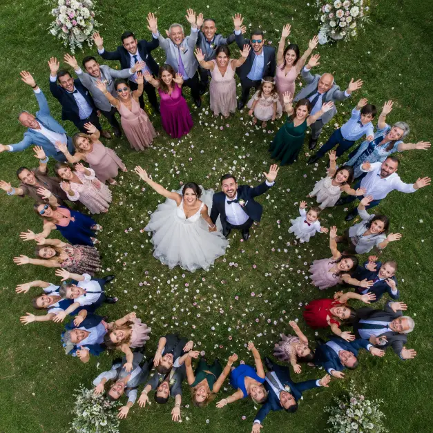 Create an overhead wedding photo of a couple standing in the center of a circle of their guests, everyone looking up toward the camera with raised arms and joyful expressions. The bride is in a white gown and the groom in a dark suit, holding hands in the middle. The guests form a loose ring around them on a grassy lawn decorated with scattered flower petals. The camera is positioned directly above in a high aerial perspective looking straight down. Bright, even daylight with vivid, celebratory tones. Shot with a wide-angle lens for the full scene. Use aspect ratio 1:1.