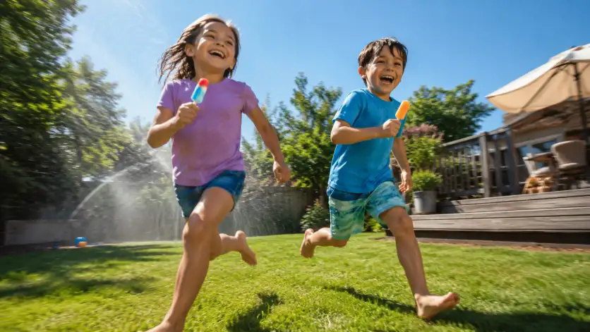Create a photo of two children around 7 years old running across a grassy backyard holding colorful popsicles, captured mid-stride with big smiles. They're wearing bright summer t-shirts, shorts, and bare feet. The camera is at a low angle near the grass, panning with their movement for a slight motion blur in the background. The background shows a sunny backyard with a sprinkler misting water, a wooden deck, and a clear blue sky. Vibrant, playful summer tones with warm natural light. Candid, joyful aesthetic. Use aspect ratio 16:9.