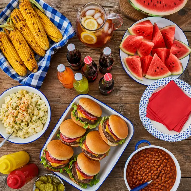 Create an overhead flat-lay photo of a full cookout spread on a rustic wooden table, including grilled corn, burgers, watermelon slices, potato salad, baked beans, and a stack of paper plates with red napkins. Glass bottles of soda and a pitcher of iced tea sit among the dishes. The camera is positioned directly above looking straight down. Bright natural daylight with vivid, saturated summer colors. Clean, appetizing food photography style. Use aspect ratio 1:1.
