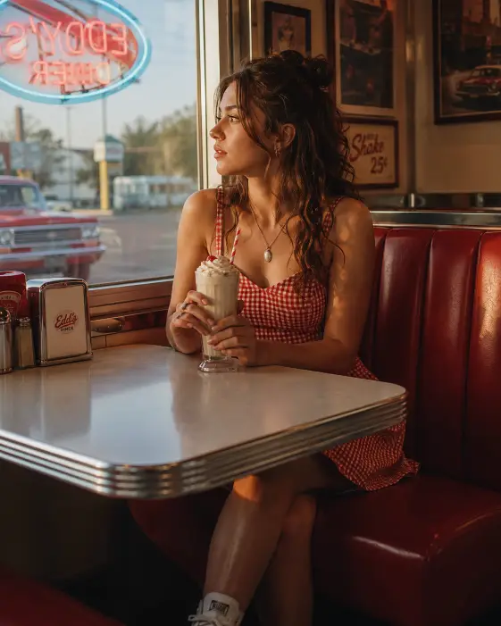 Create a photo of a young woman sitting in a vintage diner booth by the window, holding a milkshake with both hands and looking out at the parking lot. She is wearing a red gingham sundress with white sneakers and a small silver locket. Her hair is in a loose half-up style. The background shows classic diner details including red vinyl seats, chrome trim, and a neon sign reflected faintly in the window glass, with warm late afternoon light coming through. The camera is at eye level across the table, slightly tight framing. Warm nostalgic tones with soft highlights. Shot with a 50mm lens, shallow depth of field. Use aspect ratio 4:5.