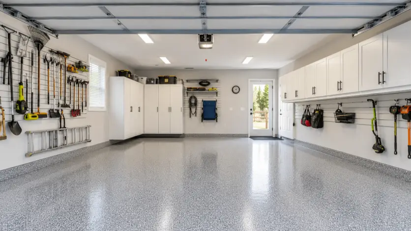 Create a real estate listing photo of a clean two-car garage with epoxy-coated gray floors, white wall-mounted storage cabinets, and neatly hung tools on a pegboard. The space is empty of cars to show its size, with good overhead lighting and a side door letting in daylight. The camera is at chest height shooting in from the open garage door to capture the full depth. Bright, even lighting keeps the space crisp and clean. Organized, functional style. Use aspect ratio 16:9.