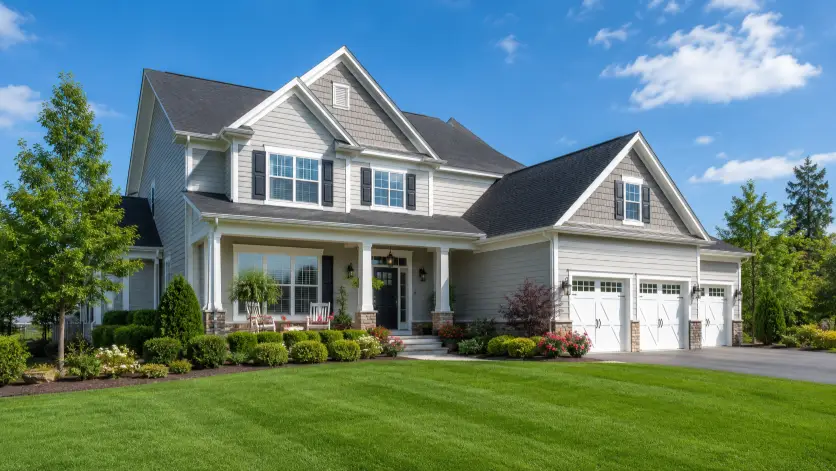 Create an exterior real estate photo of a beautiful family home shot during the day from across the front lawn, capturing the full front facade. The home has clean siding, well-kept landscaping with trimmed hedges and flower beds, a paved driveway, and a clear blue sky with a few soft clouds. The camera is positioned low and slightly angled to show the front and one side of the house for depth. Bright, warm natural light with rich green grass and crisp detail. Polished, professional listing style. Use aspect ratio 16:9.