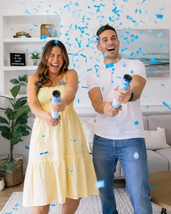 Create a photo of a couple in their living room each firing a handheld confetti cannon, sending blue streamers and confetti flying across the frame. The woman is mid-laugh with her eyes squeezed shut and the man is grinning widely. She is wearing a pastel yellow sundress and he is in a relaxed white tee with denim. The camera is at eye level capturing both of them with motion in the confetti. Bright, airy indoor lighting. Playful, high-energy mood with vivid color. Use aspect ratio 4:5.