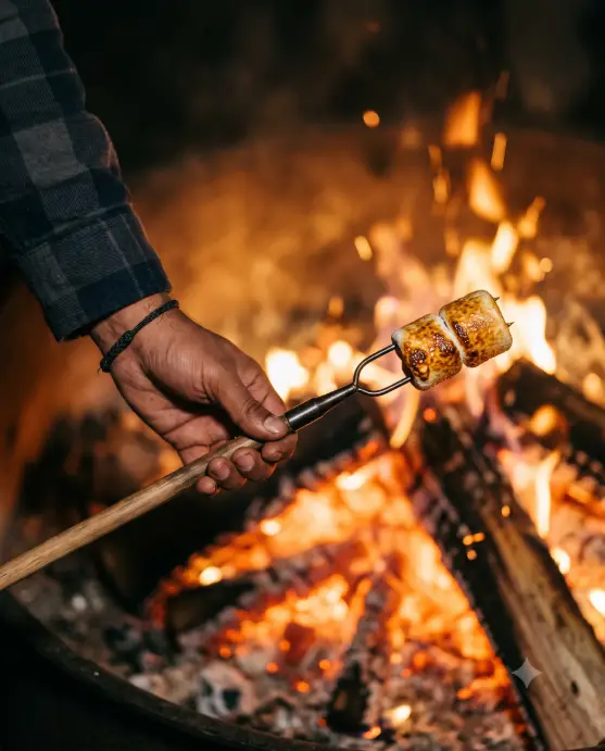 Create a close-up photo of a hand holding a long roasting stick with two golden marshmallows hovering just above the glowing embers of a campfire. The person is wearing a dark flannel sleeve pushed up to the forearm. The focus is sharp on the marshmallows and the tip of the stick, with the bright orange fire and embers blurred softly in the background. The camera is positioned low and close at eye level with the fire, with a very shallow depth of field. Warm, glowing firelight fills the frame with deep amber and red tones. Cozy, detail-focused mood. Shot with a 50mm macro lens. Use aspect ratio 4:5.