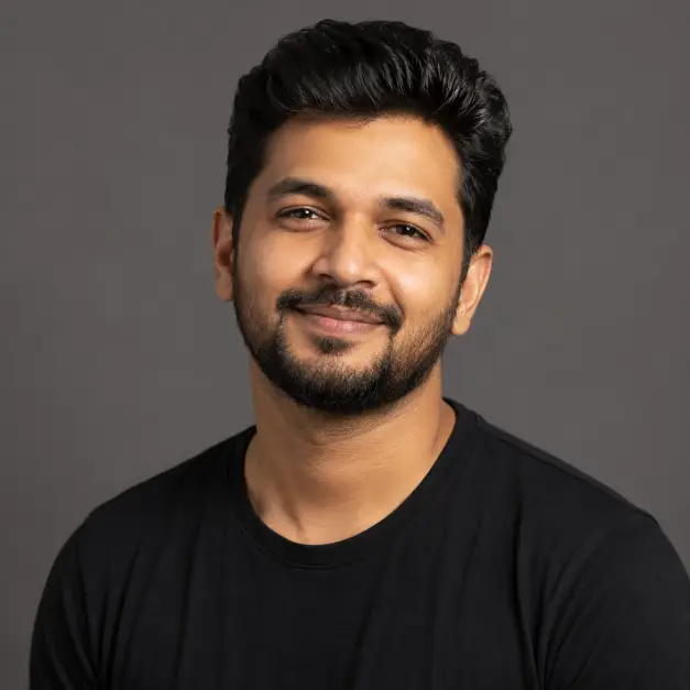 Create a professional studio headshot of a man in his early 30s, photographed from the chest up. He is facing the camera with a slight head tilt and a warm, confident smile. He wears a plain black crew-neck t-shirt with no visible logos. His hair is neatly styled and his skin looks natural with soft, even texture. Shoot at eye level with an 85mm lens look and shallow depth of field. Lighting is soft studio key light from the front left with gentle fill, no harsh shadows. The background is a smooth, solid medium-gray studio backdrop. The overall style is clean, modern, and editorial. Use aspect ratio 1:1.