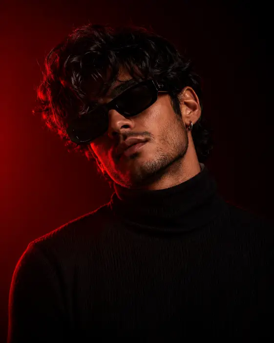 Create a dramatic studio portrait of a young man against a dark background lit with deep red gel lighting glowing from one side. He has thick wavy black hair falling over his forehead and light stubble, wearing black rectangular sunglasses, a small silver hoop earring, and a fitted black ribbed turtleneck sweater. His head is tilted slightly with his chin angled down toward one shoulder, giving a cool, confident attitude. Shoot at eye level, framed from the chest up. The red rim light traces the edge of his shoulder and hair while soft shadows keep the rest moody and dark. Rich crimson and black tones with a sleek editorial finish. Use aspect ratio 4:5.