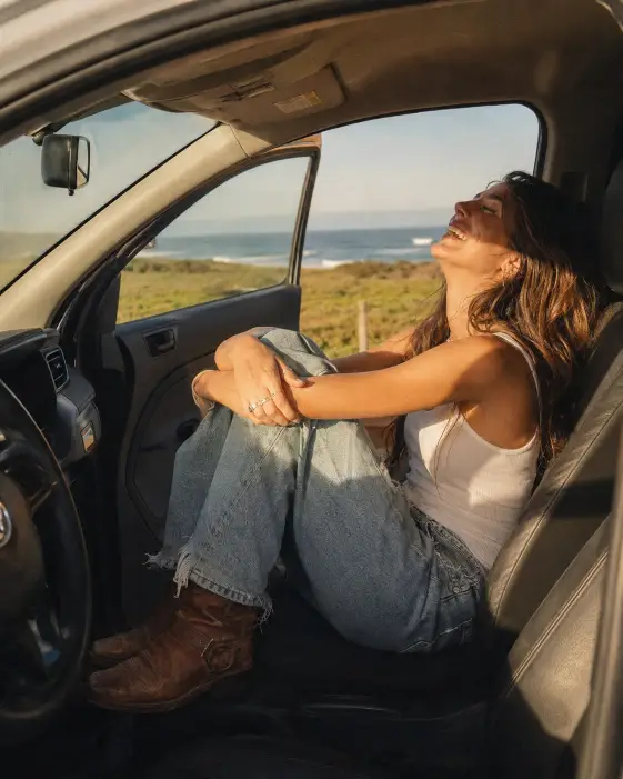 Create a candid photo of a young woman sitting sideways in the passenger seat of a car with the door open, knees pulled up toward her chest and arms crossed loosely over them. She is laughing with her eyes closed, head tilted back against the seat. She wears a white tank top, loose light-wash jeans with distressed hems, and worn brown leather boots, with several silver rings on her fingers. Warm late afternoon sunlight streams through the windshield onto her face. Through the open door and window behind her, a green coastal field stretches out toward the ocean under a soft blue sky. Shoot from just outside the driver's side at eye level with a natural, unposed feel. Warm sun-washed tones with a slight film grain. Use aspect ratio 4:5.