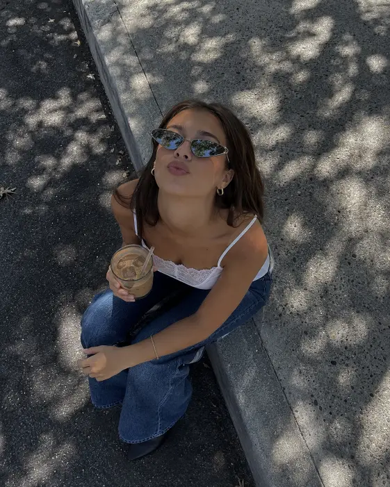 Create a photo of a young woman sitting on a shaded sidewalk curb, shot from a high overhead angle looking almost straight down at her. She is looking up at the camera with her chin raised and lips slightly pursed, wearing narrow reflective cat-eye sunglasses, a white lace-trimmed tank top, and blue jeans. One hand holds an iced drink in a clear glass. Her straight brown hair falls behind her shoulders, and she wears small gold hoop earrings and a thin bracelet. The background is textured grey asphalt and a speckled concrete sidewalk with dappled tree shade patterns across the ground. Cool, slightly desaturated street-style tones with strong texture detail. Use aspect ratio 4:5.