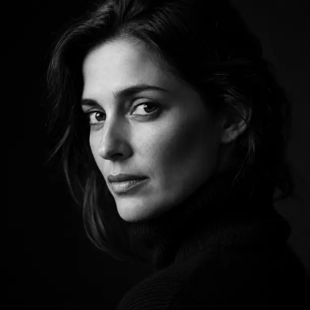 Create a dramatic black and white portrait of a woman in her 30s, framed tightly from the shoulders up. She is facing slightly away from the camera with her eyes looking back toward the lens, expression calm and intense. She wears a dark turtleneck sweater, hair loosely tucked behind one ear. Shoot at eye level with a shallow depth of field. Lighting is a single soft key light from one side, leaving the other half of her face in gentle shadow with rich contrast and deep blacks. The background is pure dark gray fading to black. The style is fine art studio portraiture with visible natural skin texture. Use aspect ratio 1:1.