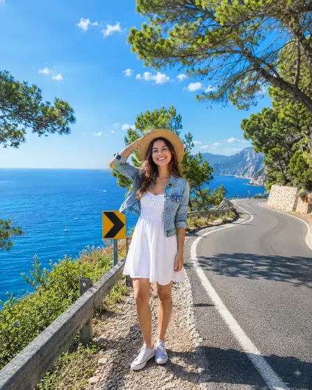 Create a photo of a young woman standing at the edge of a quiet coastal road on a bright sunny day, facing the camera with a cheerful smile, one hand holding a sun hat on her head. She is wearing a white sundress with a light denim jacket and canvas sneakers. Behind her, the road curves along a hillside with a yellow arrow sign, guardrails, and lush green trees framing a sparkling deep blue sea below. Strong summer sunlight with crisp shadows. The camera is at eye level from across the road. Vivid blues and fresh greens, bright joyful summer aesthetic, travel photography style. Use aspect ratio 4:5.
