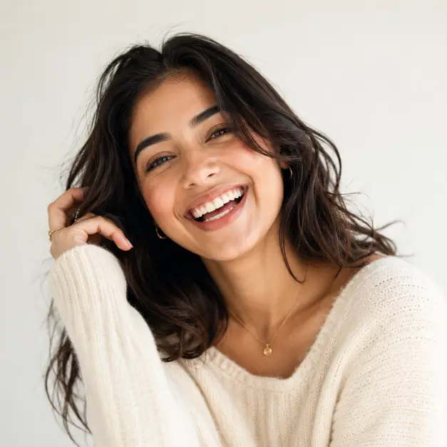 Create a cheerful portrait of a woman in her early 20s laughing naturally, framed from the chest up. Her head is tilted slightly with genuine joy in her expression, one hand lightly touching her hair. She wears a soft cream knit sweater with minimal jewelry, hair in loose natural waves with fresh, glowy makeup. Shoot at eye level with a shallow depth of field. Lighting is bright, soft, and even, like diffused daylight in a white studio. The background is a clean off-white wall with a very subtle warm tone. The overall look is bright, airy, and full of positive energy. Use aspect ratio 1:1.