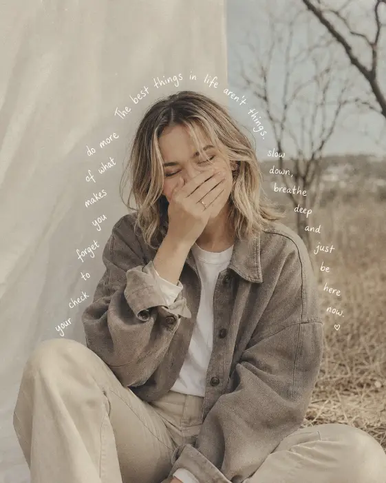 Create a soft, faded lifestyle photo of a young woman sitting outdoors and laughing shyly with one hand covering her mouth, eyes closed. She has shoulder-length blonde hair with darker roots and wears an oversized taupe shacket over a white t-shirt with matching light trousers. The background shows a white fabric backdrop on one side and blurred bare trees with dry grass on the other, in washed-out beige and grey tones. Shoot at eye level with bright, diffused daylight. Add a handwritten-style white text overlay that curves in an arc around her head and shoulders, following the shape of her silhouette, as if a favorite quote is written around her. Muted, dusty, vintage-filtered color grading. Use aspect ratio 4:5.
