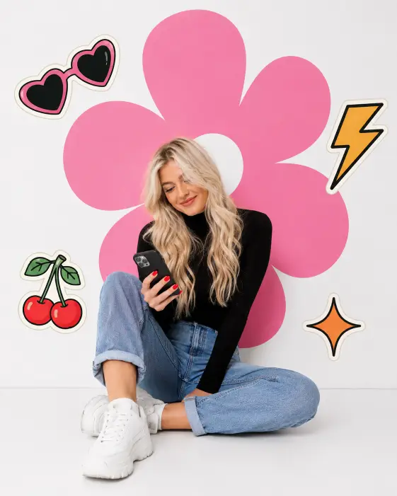 Create a clean studio photo of a young woman with long wavy platinum blonde hair sitting on a white floor against a plain white background, one knee bent up and the other leg tucked under. She is looking down at a smartphone held in one hand with a soft smile. She wears a fitted black turtleneck, cuffed light blue jeans, and chunky white sneakers, with red painted nails. Behind her, add a large flat pink flower graphic with rounded petals centered on the wall like a designed backdrop. Scatter a few retro sticker-style illustrations around her: heart-shaped sunglasses, cherries, a lightning bolt, and an orange sparkle. Bright even studio lighting with a playful, editorial-graphic look. Use aspect ratio 4:5.