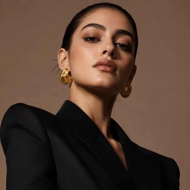 Create a high-fashion editorial portrait of a woman in her 20s, framed from the shoulders up. She is posed with her chin slightly raised and shoulders angled, gazing at the camera with a strong, confident expression. She wears a structured black blazer with sharp shoulders and bold gold earrings, hair slicked back, makeup featuring defined brows and a subtle smoky eye. Shoot slightly below eye level for a powerful feel, with a shallow depth of field. Lighting is crisp studio beauty lighting with a soft shadow under the jaw. The background is a solid warm taupe studio backdrop. The style is sharp, editorial, and fashion-forward. Use aspect ratio 1:1.