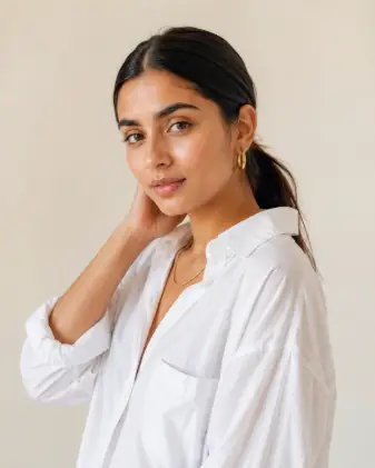 Create a portrait of a young woman wearing an oversized crisp white button-down shirt with the top two buttons open and sleeves rolled to the elbow. Her hair is in a sleek center-parted low ponytail, and she wears thin gold hoop earrings and a delicate gold chain necklace. She is standing with one hand gently touching the side of her neck, body angled slightly away while she looks back at the camera with a soft, confident gaze. Shoot at eye level, slightly close-up, with a shallow depth of field. The lighting is bright and airy, soft morning light. The background is a clean cream-colored studio backdrop. Neutral tones, fresh and minimal. Editorial lifestyle style. Use aspect ratio 4:5.
