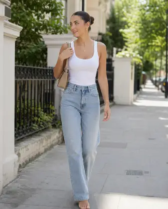 Create a candid street style photo of a young woman walking along a quiet city sidewalk on a bright day. She is wearing a fitted white tank top tucked into straight-leg light-wash jeans, with a small structured beige shoulder bag. Her hair is in a slicked-back bun and she wears gold hoops and minimal jewelry. She is captured mid-stride with a relaxed walk, one hand adjusting her bag strap, looking slightly off-camera with a soft natural smile. Shoot at eye level from a short distance with a shallow depth of field. The lighting is bright, soft daylight. The background is a clean, blurred street with pale buildings and greenery. Neutral tones, effortless lifestyle aesthetic. Use aspect ratio 4:5.