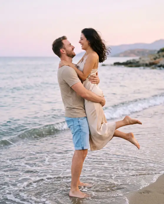 Create a photo of a man lifting a woman up in the air at the edge of the sea, holding her at his chest height while they both laugh and look into each other's eyes. He is wearing a fitted beige t-shirt and rolled light-blue denim shorts, standing barefoot in shallow water, and she is in a champagne satin slip dress with her bare feet kicked up behind her and her hair blowing in the breeze. The background is a calm pastel ocean and a pale pink and lavender evening sky. The camera is at waist height, shooting from the side to capture both profiles. Soft, airy, film-inspired tones. Shot with an 85mm lens, creamy background blur. Use aspect ratio 4:5.