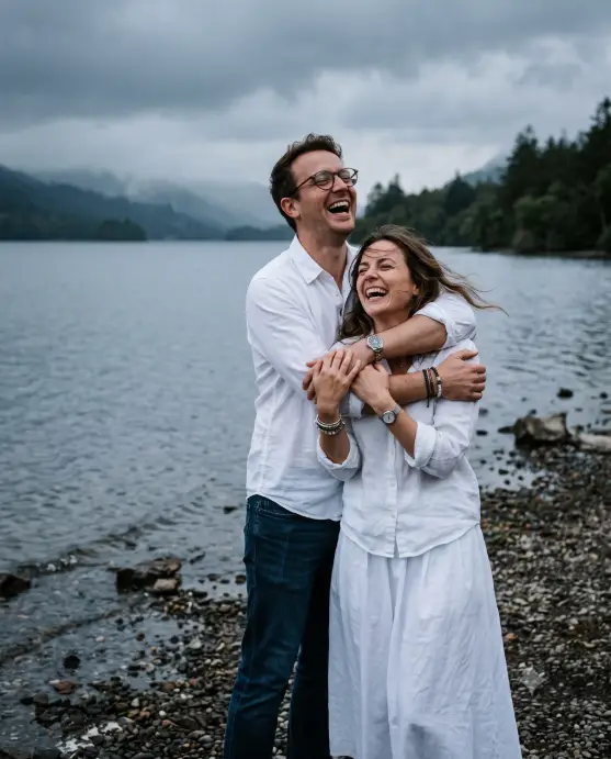 Create a candid photo of a couple on a pebbly lakeshore on an overcast evening, the man hugging the woman tightly from behind with his arms wrapped over her shoulders, both laughing hard with heads tilted back. He is wearing round glasses, a loose white cotton shirt, and dark jeans, and she is in a relaxed white blouse and flowing white skirt with a silver watch and bracelets on her wrists. The background shows a calm gray-blue lake fading into misty trees along the shore. The camera is at eye level with a slight tilt, capturing them off-center. Moody, muted tones with soft natural light and a slightly stormy sky. Shot with a 35mm lens, documentary style. Use aspect ratio 4:5.