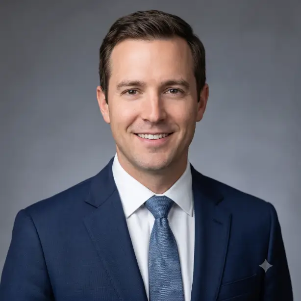 Create a professional corporate headshot of the person in the uploaded photo, keeping their face and features exactly the same. They are wearing a well-fitted navy blue suit jacket over a crisp white dress shirt, shoulders relaxed and squared to the camera with a confident, approachable smile. Shoot at eye level from the chest up. Soft studio lighting with a key light from the front left and gentle fill, creating even skin tones with no harsh shadows. The background is a smooth, softly blurred medium gray studio backdrop. Clean, sharp, editorial corporate photography style. Use aspect ratio 1:1.