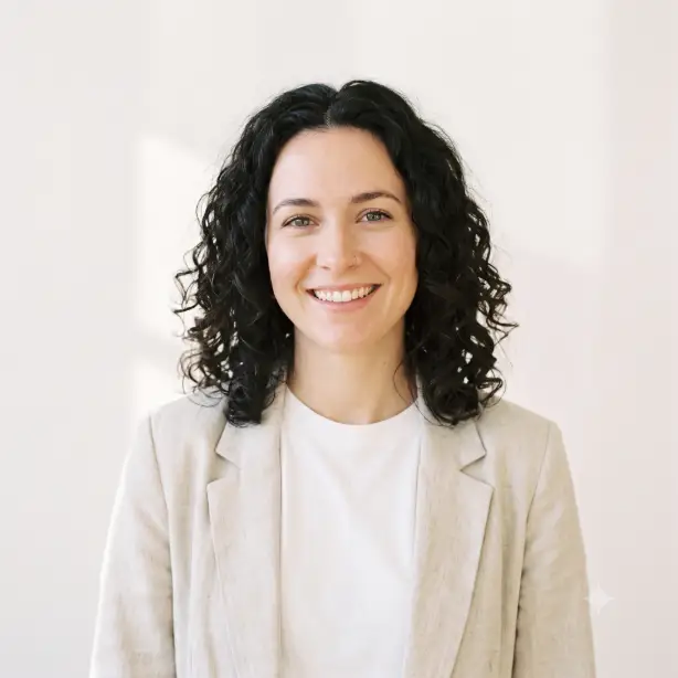 Create a minimalist professional headshot of the person in the uploaded photo, keeping their features exactly the same. They are wearing a plain white crew neck t-shirt under a light beige unstructured blazer, standing relaxed with a soft, genuine smile and direct eye contact. Shoot at eye level, framed from the shoulders up. Bright, diffused natural daylight from a large window in front, creating luminous even skin. The background is a plain warm white wall with a faint soft shadow. Airy, clean, Scandinavian-style editorial photography with light neutral tones. Use aspect ratio 1:1.