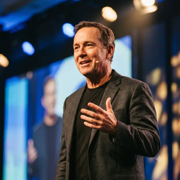 Create a dynamic professional portrait of the person in the uploaded photo, keeping their face identical. They are wearing a smart dark blazer over a plain black t-shirt with a thin lapel microphone clipped on, mid-gesture with one hand raised naturally as if making a point, expression animated and engaging with a warm smile. Shoot at a slight three-quarter angle at eye level, framed from the chest up. Warm stage-style lighting from the front with a soft blue ambient glow behind. The background is a blurred conference stage with bokeh lights in deep blue and gold. Energetic event photography style with rich, warm contrast. Use aspect ratio 1:1.