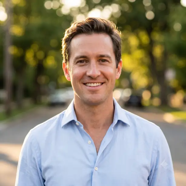 Create an outdoor professional headshot of the person in the uploaded photo, keeping their features identical. They are wearing a light blue button-down shirt with the top button open, standing relaxed with a warm, open smile and eyes engaged with the camera. Shoot at eye level, framed from the shoulders up with shallow depth of field. Golden hour sunlight from behind creating a soft rim light on the hair, with reflected warm fill light on the face. The background is a blurred tree-lined street with soft green and gold bokeh. Natural, candid lifestyle photography style. Use aspect ratio 1:1.