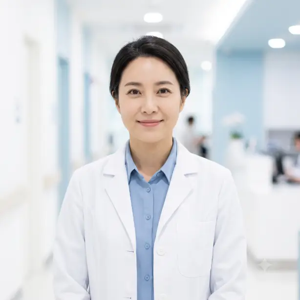 Create a professional headshot of the person in the uploaded photo, preserving their likeness. They are wearing a clean white medical coat over a soft blue shirt, standing upright with a gentle, reassuring smile and kind eyes directed at the camera. Shoot at eye level from the chest up. Bright, soft, clinical-clean lighting with no harsh shadows. The background is a softly blurred bright clinic hallway in whites and pale blues. Trustworthy, warm professional photography style with fresh, airy tones. Use aspect ratio 1:1.
