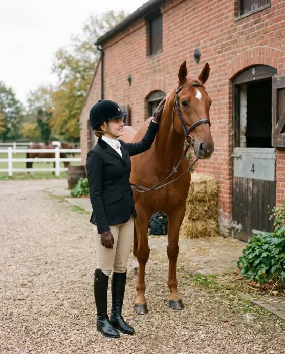Create a photo of a young woman standing beside a chestnut horse outside a traditional brick stable, one hand gently resting on the horse's neck. She is wearing a fitted black riding jacket, a white show shirt, tan breeches, tall polished black leather riding boots, and thin leather gloves. Her hair is tucked neatly under a black velvet riding helmet. The camera is at eye level with a medium framing that includes both her and the horse's head. Soft overcast daylight gives even, flattering light, and the background shows stable doors, hay bales, and a white wooden fence. Classic equestrian editorial style with muted earth tones. Use aspect ratio 4:5.