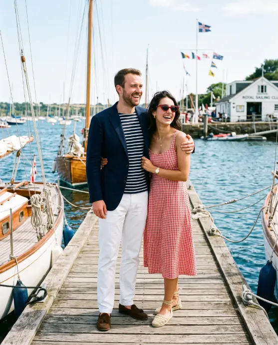 Create a photo of a couple standing together on a weathered wooden dock at a sailing club, the man standing slightly behind the woman with one arm around her shoulder. He is wearing a navy blazer over a striped Breton shirt and white trousers, and she is wearing a red and white gingham sundress with espadrilles and cat-eye sunglasses pushed into her hair. Both are laughing naturally, looking off toward the water. The camera is at eye level with medium framing. Bright, crisp midday coastal light, with moored wooden sailboats, rippling blue water, and distant club flags in the background. Nautical editorial style with navy, white, and red tones. Use aspect ratio 4:5.