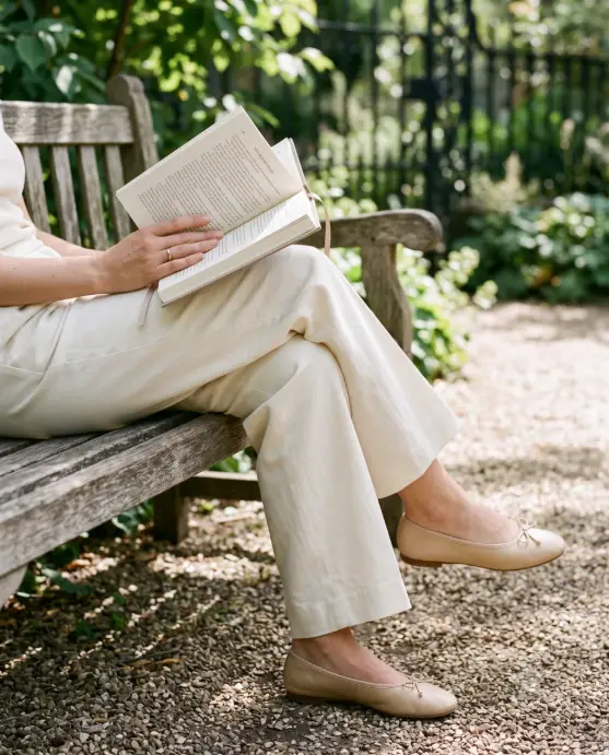 Create a photo of a woman sitting on a classic wooden park bench, cropped from the waist down to focus on her crossed legs and the open hardcover book resting on her lap. She is wearing pressed wide-leg cream trousers and buttery beige leather ballet flats, with one hand holding the page and a thin gold ring visible. Shoot at a slight downward angle from bench height with an 85mm lens and soft background blur. Dappled late-morning light filters through green leaves onto the gravel path, with a blurred wrought-iron fence behind. Palette of cream, beige, and soft green. The mood is calm, detailed, and quietly polished. Use aspect ratio 4:5.