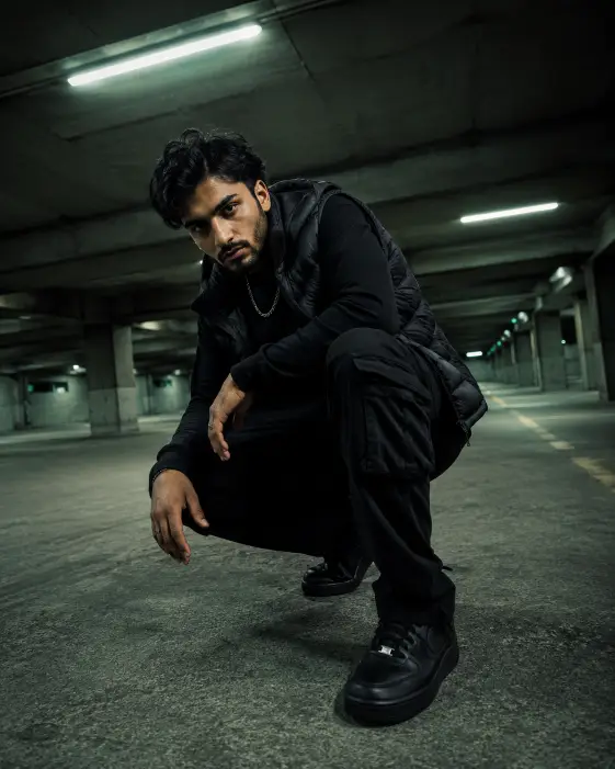 Create a photo of a man crouching in an empty concrete parking garage at night, one forearm resting on his knee and his gaze locked on the camera with an intense expression. He is wearing an all-black outfit with a puffer vest over a long-sleeve shirt, cargo pants, and black sneakers, plus a silver chain. Rows of fluorescent ceiling lights stretch behind him, creating strong leading lines and greenish-white highlights against deep shadows. The camera is low, near ground level, shooting slightly upward. Gritty, high-contrast, editorial streetwear mood. Shot with a 24mm wide-angle lens. Use aspect ratio 4:5.