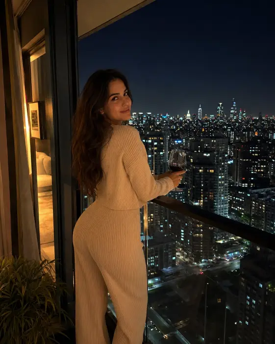 Create a photo of a woman standing on a high-rise apartment balcony at night, leaning gently on the glass railing and looking back at the camera over her shoulder. She is wearing a ribbed knit lounge set in cream with her hair down in soft waves, holding a glass of wine in one hand. Behind and below her, an endless city skyline glitters with lights under a starless deep blue sky. Warm light from the apartment interior spills onto her from the side. The camera is at eye level from inside the balcony doorway. Cozy, intimate mood with warm and cool tone contrast. Shot with a 35mm lens. Use aspect ratio 4:5.