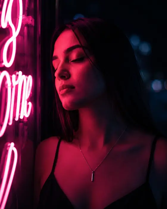 Create a close-up portrait of a young woman standing beside a glowing pink neon sign on a dark city wall at night, her face turned toward the light with her eyes gently closed. She is wearing a black slip dress with a thin silver necklace, and her sleek straight hair falls past her shoulders. The pink neon light washes over one side of her face while the other side falls into soft blue shadow. The background is nearly black with faint distant city bokeh. The camera is at eye level, tight framing from the shoulders up. Bold pink and blue duotone lighting, editorial and artistic. Shot with an 85mm lens. Use aspect ratio 4:5.