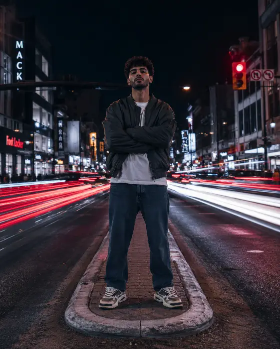 Create a photo of a stylish man standing still in the middle of a pedestrian island on a busy city street at night, arms crossed and looking directly into the camera with a confident expression. He is wearing an oversized bomber jacket, a plain white tee, straight-leg jeans, and chunky sneakers. Behind him, red and white car light trails streak across the frame from long exposure, with illuminated storefronts and traffic signals in the background. The camera is at waist height, shooting slightly upward. Sharp focus on him with motion blur everywhere else. Cool tones with pops of red and amber. Shot with a 35mm lens. Use aspect ratio 4:5.