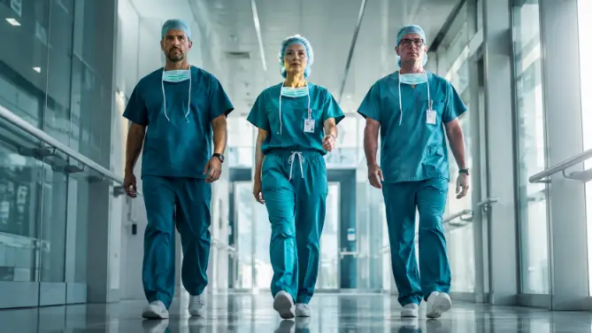 Create a photo of a team of three surgeons walking side by side down a bright hospital corridor toward the camera, captured mid-stride. The group includes a woman in the center wearing teal scrubs and a surgical cap, flanked by two male colleagues in matching scrubs with masks hanging around their necks. Their expressions are focused and determined. Shoot from a low angle at knee height to make the group look powerful. The background shows a long corridor with glass panels and soft daylight streaming in. Cinematic editorial style with cool tones and slight motion energy. Use aspect ratio 16:9.