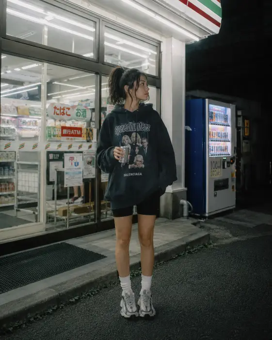 Create a photo of a young woman standing outside a brightly lit convenience store at night, holding a cold drink can and looking off to the side with a casual expression. She is wearing an oversized graphic hoodie, bike shorts, crew socks, and chunky sneakers, with her hair in a messy high ponytail. The store's fluorescent white light spills onto the sidewalk, contrasting with the dark blue night around the edges of the frame. A vending machine glows softly beside the entrance. The camera is at eye level, straight on. Cool fluorescent tones with a slightly grainy, film-like quality. Shot with a 35mm lens. Use aspect ratio 4:5.
